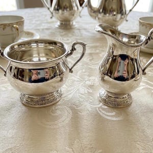 Puede incluir: Un azucarero de plata pulida con dos asas ornamentadas y una jarra de leche de plata a juego con asa curva y pico vertedor. Ambas piezas presentan una base circular con un borde floral en relieve y un acabado met&aacute;lico brillante.