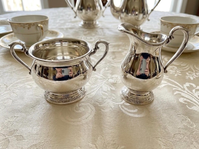 Puede incluir: Un azucarero de plata pulida con dos asas ornamentadas y una jarra de leche de plata a juego con asa curva y pico vertedor. Ambas piezas presentan una base circular con un borde floral en relieve y un acabado met&aacute;lico brillante.