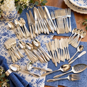 May include: A large silver flatware set featuring ornate floral detailing on the handles. The collection includes various sizes of forks, knives, and spoons, along with a ladle, a slotted serving spoon, and a serving fork, all arranged on a blue cloth.