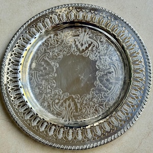 May include: A round silver-toned metal serving tray featuring an intricate openwork lattice border with leaf motifs and a central circular area decorated with embossed scrollwork and filigree patterns along the inner rim.