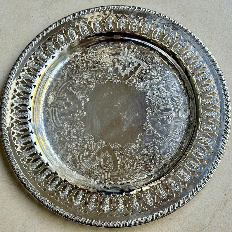 May include: A round silver-toned metal serving tray featuring an intricate openwork lattice border with leaf motifs and a central circular area decorated with embossed scrollwork and filigree patterns along the inner rim.