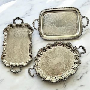 May include: Three silver-coloured serving trays with ornate, embossed floral patterns. The trays vary in shape: rectangular, oval, and a scalloped edge design. Each tray has decorative handles.