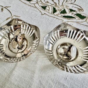 May include: Two silver-toned, decorative dishes with a floral design in the center. The dishes have a scalloped edge with vertical cutouts. One dish has a ring with a white stone.