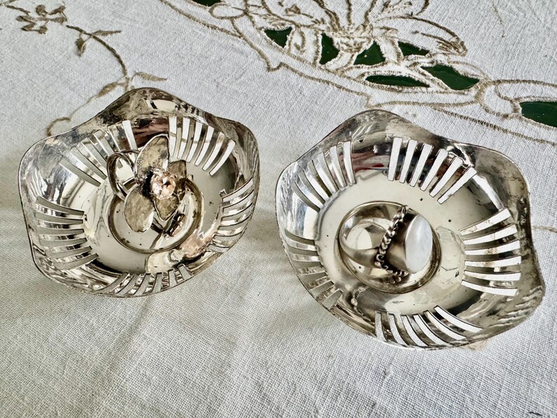 May include: Two silver-toned, decorative dishes with a floral design in the center. The dishes have a scalloped edge with vertical cutouts. One dish has a ring with a white stone.