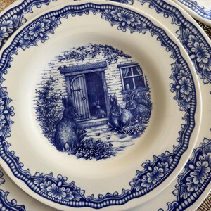 K&ouml;nnte beinhalten: Ein Set aus blauen und wei&szlig;en Keramik-Dinnertellern mit floralem Rand und einer Szene mit Kaninchen vor einem H&auml;uschen.