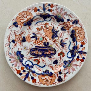 Pu&ograve; includere: Un piatto rotondo in ceramica bianca con un'urna decorativa blu scuro al centro, riempita con motivi floreali arancioni e blu. Il bordo e la superficie sono decorati con complessi motivi botanici in arancione, blu e rosa chiaro.