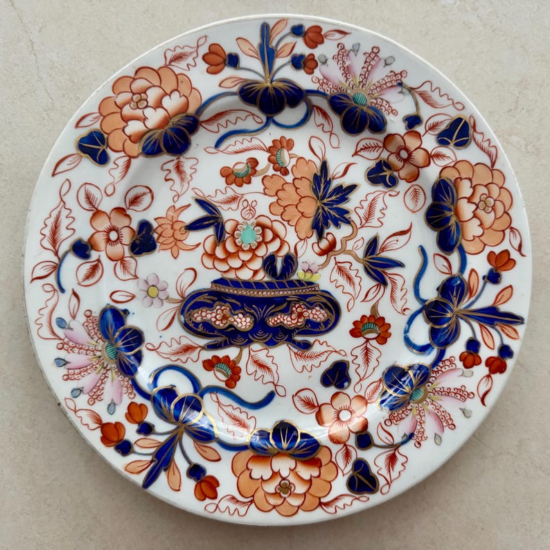 Pu&ograve; includere: Un piatto rotondo in ceramica bianca con un'urna decorativa blu scuro al centro, riempita con motivi floreali arancioni e blu. Il bordo e la superficie sono decorati con complessi motivi botanici in arancione, blu e rosa chiaro.