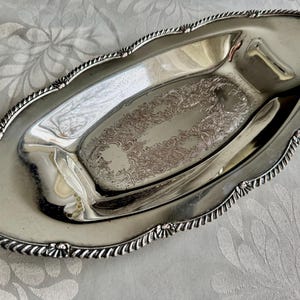 K&ouml;nnte beinhalten: Ein ovales silberfarbenes Serviertablett mit einer polierten, reflektierenden Oberfl&auml;che und einem zentralen Bereich, der mit komplizierten, gepr&auml;gten Blumenranken verziert ist. Der Rand ist mit einem erhabenen, gedrehten Seilmuster und kleinen muschelartigen Akzenten versehen.