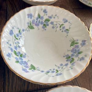 May include: White porcelain bowls with a delicate blue floral pattern and gold trim. The bowls have a scalloped edge and are arranged on a wooden surface. The floral design features small blue flowers and green leaves.