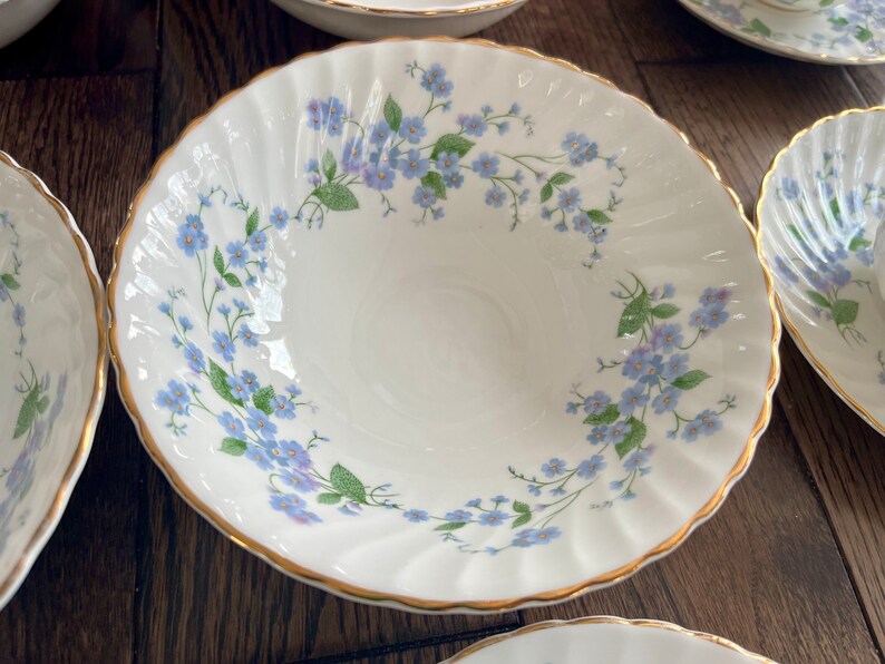 May include: White porcelain bowls with a delicate blue floral pattern and gold trim. The bowls have a scalloped edge and are arranged on a wooden surface. The floral design features small blue flowers and green leaves.