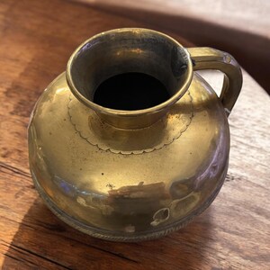 K&ouml;nnte beinhalten: Eine Messingkanne mit rundem K&ouml;rper und einem einzigen Henkel. Die Kanne hat einen dekorativen gewellten Rand und einen strukturierten Boden.