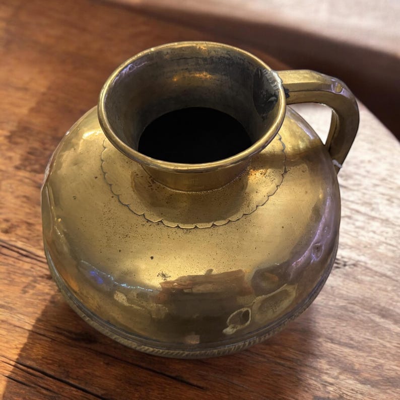 K&ouml;nnte beinhalten: Eine Messingkanne mit rundem K&ouml;rper und einem einzigen Henkel. Die Kanne hat einen dekorativen gewellten Rand und einen strukturierten Boden.