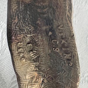 May include: Close-up of a silver-toned metal object with etched patterns and the text "VIK-INC C9" and "STERLING". The object has a textured surface with intricate designs, possibly a piece of vintage silverware.