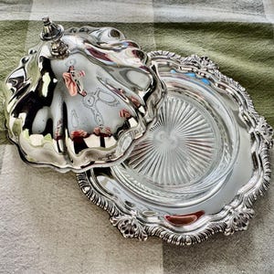 May include: A silver-plated butter dish with a lid, featuring a decorative, ornate design. The dish has a scalloped edge and a clear glass insert. The lid is open, revealing the interior. The dish is on a checkered tablecloth.