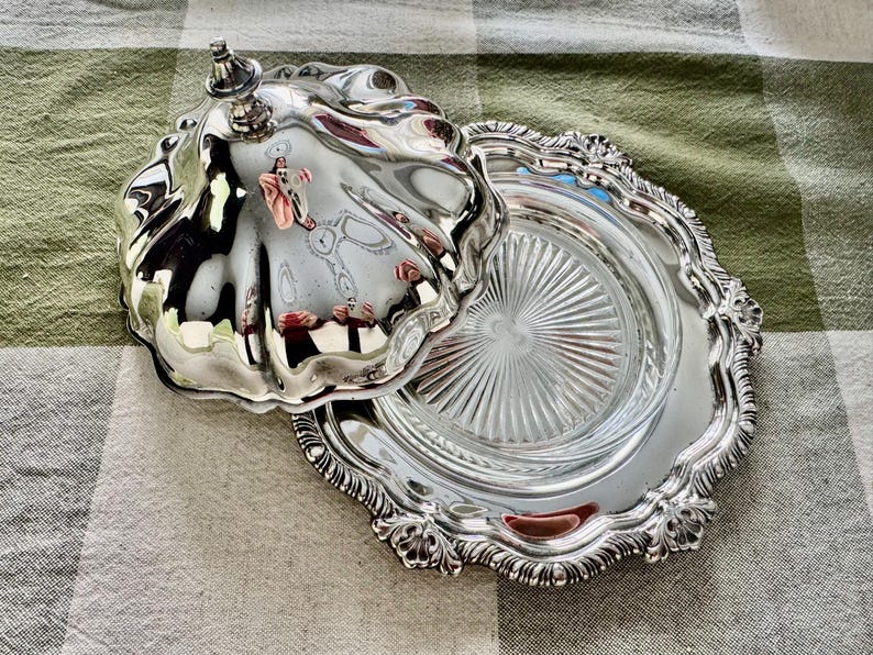 May include: A silver-plated butter dish with a lid, featuring a decorative, ornate design. The dish has a scalloped edge and a clear glass insert. The lid is open, revealing the interior. The dish is on a checkered tablecloth.