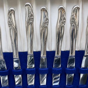 May include: A set of seven silver-toned dinner knives arranged vertically. Each knife features a polished metallic finish and a decorative handle with an embossed floral motif. The knives are held in place by blue fabric slots within a storage case.