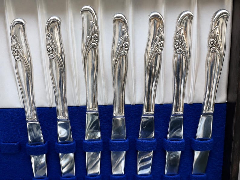 May include: A set of seven silver-toned dinner knives arranged vertically. Each knife features a polished metallic finish and a decorative handle with an embossed floral motif. The knives are held in place by blue fabric slots within a storage case.