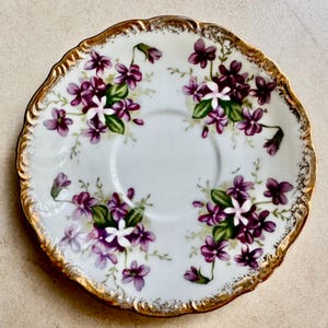 May include: A round porcelain saucer featuring a white base decorated with clusters of purple violet flowers and green leaves. The scalloped rim is finished with a decorative gold gilded edge that shows slight wear along the textured border.