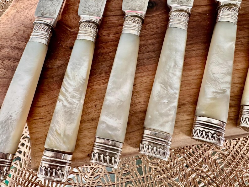 Pu&ograve; includere: Set di sei coltelli d'argento con manici in madreperla. I coltelli hanno dettagli in argento decorativi e sono disposti su una superficie in legno.