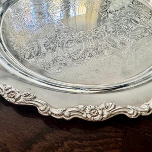 May include: A round silver serving tray featuring an intricate embossed floral and scroll pattern in the center. The edge of the tray has a scalloped border with raised flower motifs and a polished, reflective metallic finish throughout.
