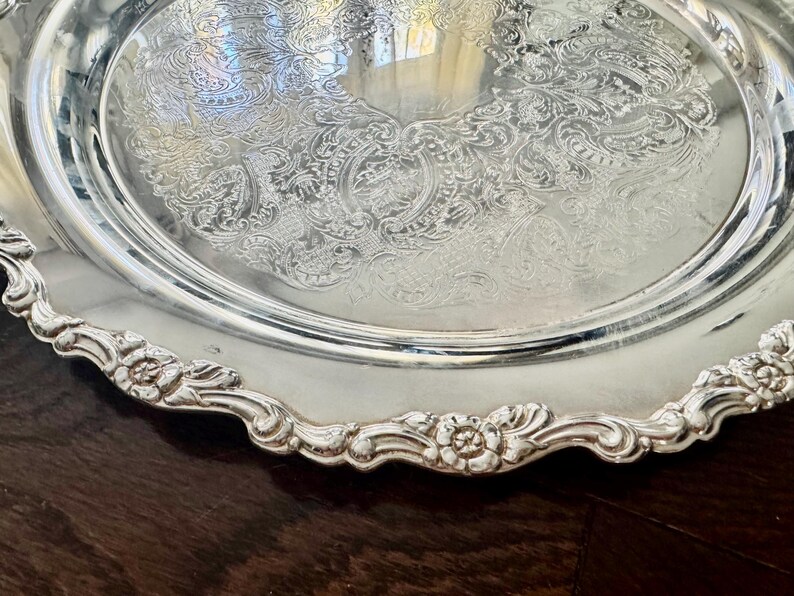 May include: A round silver serving tray featuring an intricate embossed floral and scroll pattern in the center. The edge of the tray has a scalloped border with raised flower motifs and a polished, reflective metallic finish throughout.