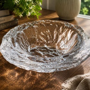 May include: A clear glass bowl with a textured, uneven surface that mimics the appearance of melting ice or flowing water. The bowl has a wide, irregular rim and a deep, rounded basin, reflecting light across its multifaceted, crystalline structure.