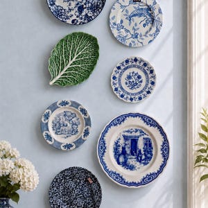 Puede incluir: Una colecci&oacute;n de siete platos decorativos expuestos en una pared, incluyendo seis platos redondos de cer&aacute;mica con motivos azules y blancos florales, paisaj&iacute;sticos y animales, y un plato verde con forma de hoja con nervaduras detalladas.