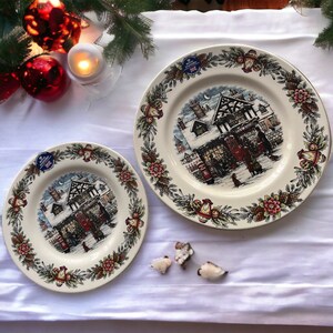 May include: Two cream-colored ceramic plates featuring a central illustration of a snow-covered village scene with a toy shop, surrounded by a border of pine cones, holly, and lanterns. Each plate bears a Royal Stafford sticker.