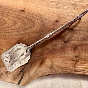 May include: Silver-plated serving tongs with an ornate design on the handles and a decorative, scalloped edge on the serving end. The tongs are resting on a wooden surface.