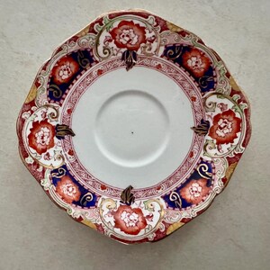 Pu&ograve; includere: Un piattino in ceramica rotondo con bordo smerlato, centro bianco e bordo decorativo. Il bordo presenta un motivo di fiori rossi, pannelli blu scuro e dettagli a volute dorate disposti in un design simmetrico.