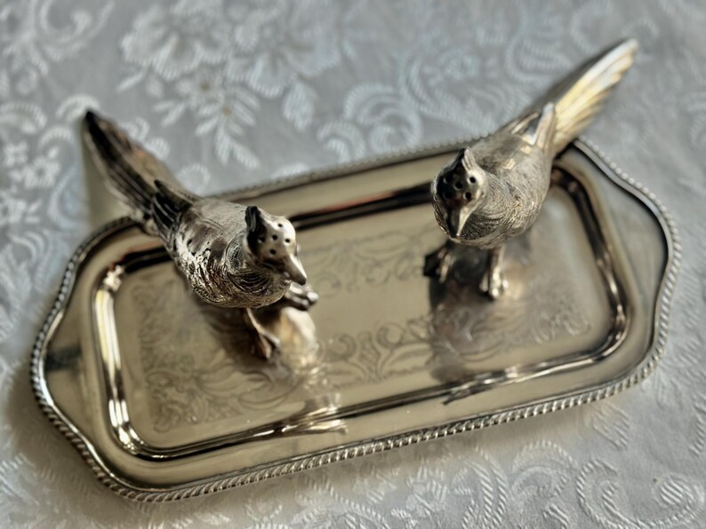 May include: A silver-toned serving tray with two bird-shaped salt and pepper shakers. The tray has a rectangular shape with a decorative edge and a floral pattern. The bird shakers have detailed feather and eye features.