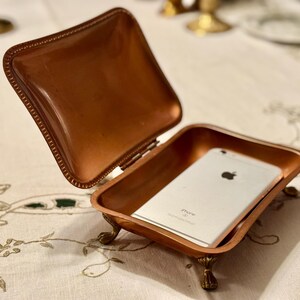 May include: A small, open copper-colored box with a hinged lid, resting on ornate feet. Inside the box is a silver smartphone. The box is on a white tablecloth with a floral design. The lid is open.