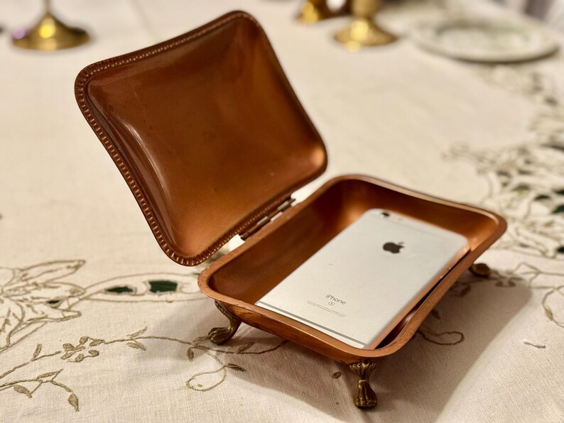 May include: A small, open copper-colored box with a hinged lid, resting on ornate feet. Inside the box is a silver smartphone. The box is on a white tablecloth with a floral design. The lid is open.