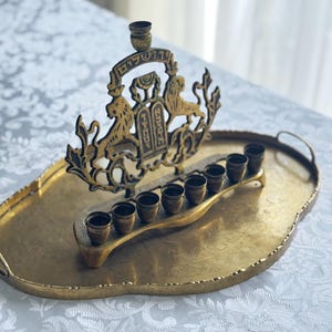 May include: A gold-toned menorah with a decorative crest featuring lions and Hebrew text, set on a matching oval tray. The menorah has eight candle holders and a central holder. The tray has handles and a textured surface.