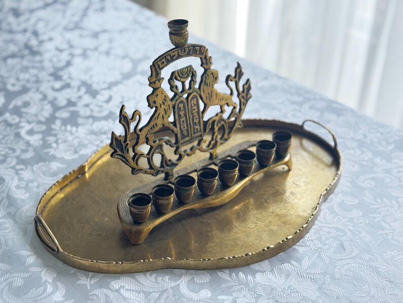 May include: A gold-toned menorah with a decorative crest featuring lions and Hebrew text, set on a matching oval tray. The menorah has eight candle holders and a central holder. The tray has handles and a textured surface.