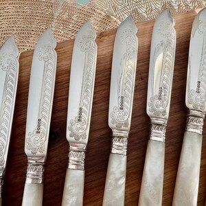 Pu&ograve; includere: Set di otto coltelli da burro in argento con manici in madreperla. Ogni coltello ha un design elaborato e la parola "Boggs" incisa sulla lama.