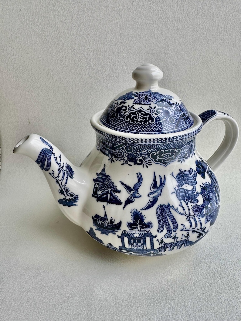 K&ouml;nnte beinhalten: Eine wei&szlig;e Teekanne mit einem blauen und wei&szlig;en Weidenmuster. Die Teekanne hat einen runden K&ouml;rper und einen Deckel mit einem Knopf oben. Die Teekanne ist mit einem traditionellen chinesischen Design verziert.