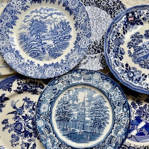 May include: A collection of six ceramic plates featuring blue and white transferware patterns. The designs include detailed landscapes, architectural structures, floral borders, and bird motifs on cream-colored backgrounds with scalloped edges.