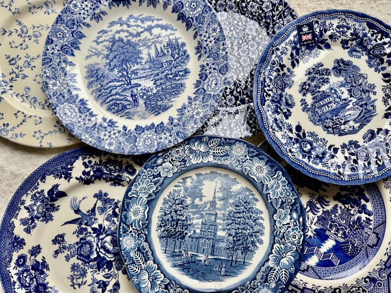 May include: A collection of six ceramic plates featuring blue and white transferware patterns. The designs include detailed landscapes, architectural structures, floral borders, and bird motifs on cream-colored backgrounds with scalloped edges.