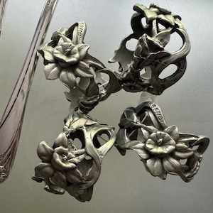 Pu&ograve; includere: Un set di tre portatovaglioli in metallo color argento con un design floreale in rilievo raffigurante narcisi, completi di steli e foglie dettagliati. Ogni anello presenta una fascia circolare e un marchio impresso sul bordo interno.