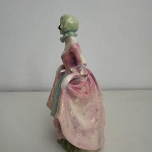 以下が含まれることがあります： 白い花柄のピンクのドレス、水色のアンダースカート、淡い緑色のヘッドカバーを身につけた人物の磁器製フィギュア。横向きに立っており、片手で長く流れるようなスカートの端を掴んでいる様子が表現されている。