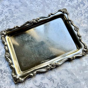 Puede incluir: Una bandeja de servicio rectangular de metal plateado con un borde elevado y ornamentado que presenta intrincados adornos de volutas y motivos de conchas. La superficie central es lisa y reflectante, con un sutil dise&ntilde;o floral grabado.