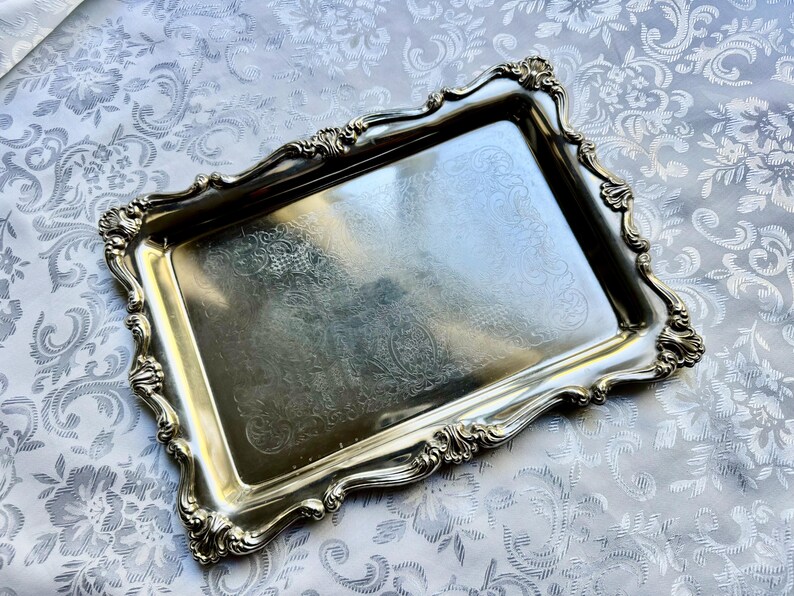 Puede incluir: Una bandeja de servicio rectangular de metal plateado con un borde elevado y ornamentado que presenta intrincados adornos de volutas y motivos de conchas. La superficie central es lisa y reflectante, con un sutil dise&ntilde;o floral grabado.