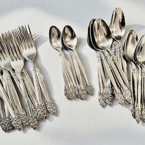 May include: A collection of vintage silverware, including forks and spoons, arranged on a white surface. The silverware has ornate, detailed handles and a shiny, reflective finish. The forks and spoons are grouped together, suggesting a complete set for dining.