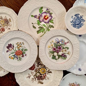 May include: A collection of assorted ceramic plates and bowls featuring various floral patterns, including hibiscus, tulips, and mixed bouquets. The pieces have white embossed rims with intricate textures and scalloped edges, showcasing diverse botanical designs.