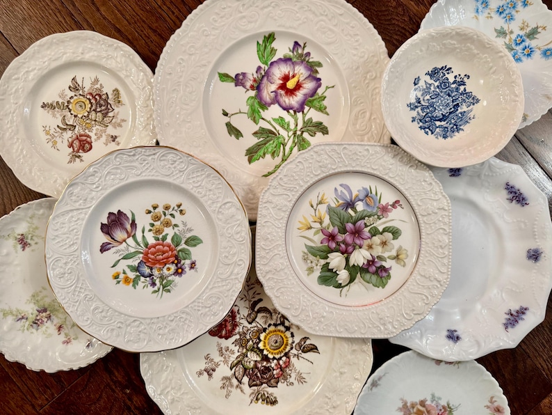 May include: A collection of assorted ceramic plates and bowls featuring various floral patterns, including hibiscus, tulips, and mixed bouquets. The pieces have white embossed rims with intricate textures and scalloped edges, showcasing diverse botanical designs.