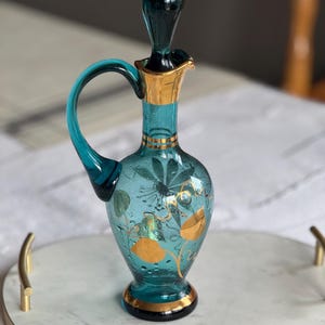 May include: A teal blue glass bottle with a gold stopper and gold painted floral designs. The bottle has a round body and a curved handle. It is sitting on a white marble tray with a gold rim.