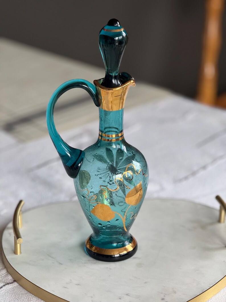 May include: A teal blue glass bottle with a gold stopper and gold painted floral designs. The bottle has a round body and a curved handle. It is sitting on a white marble tray with a gold rim.