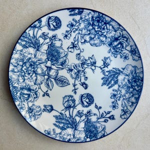 May include: A round ceramic plate featuring a white base with a detailed blue floral pattern. The design consists of various blooming flowers, buds, and leaves arranged across the surface, finished with a thin blue rim around the edge.