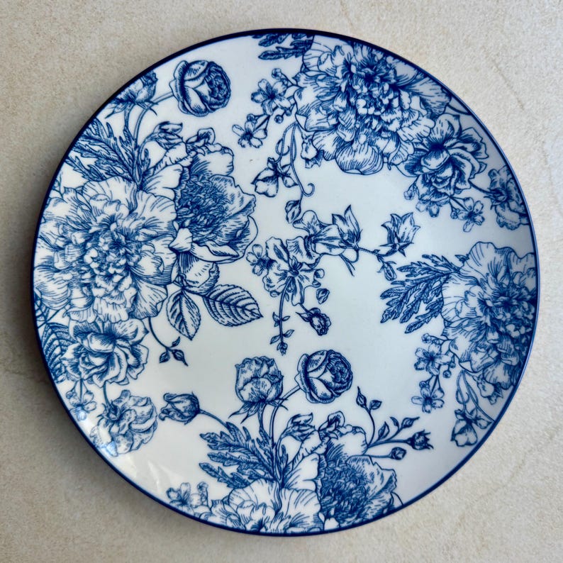 May include: A round ceramic plate featuring a white base with a detailed blue floral pattern. The design consists of various blooming flowers, buds, and leaves arranged across the surface, finished with a thin blue rim around the edge.
